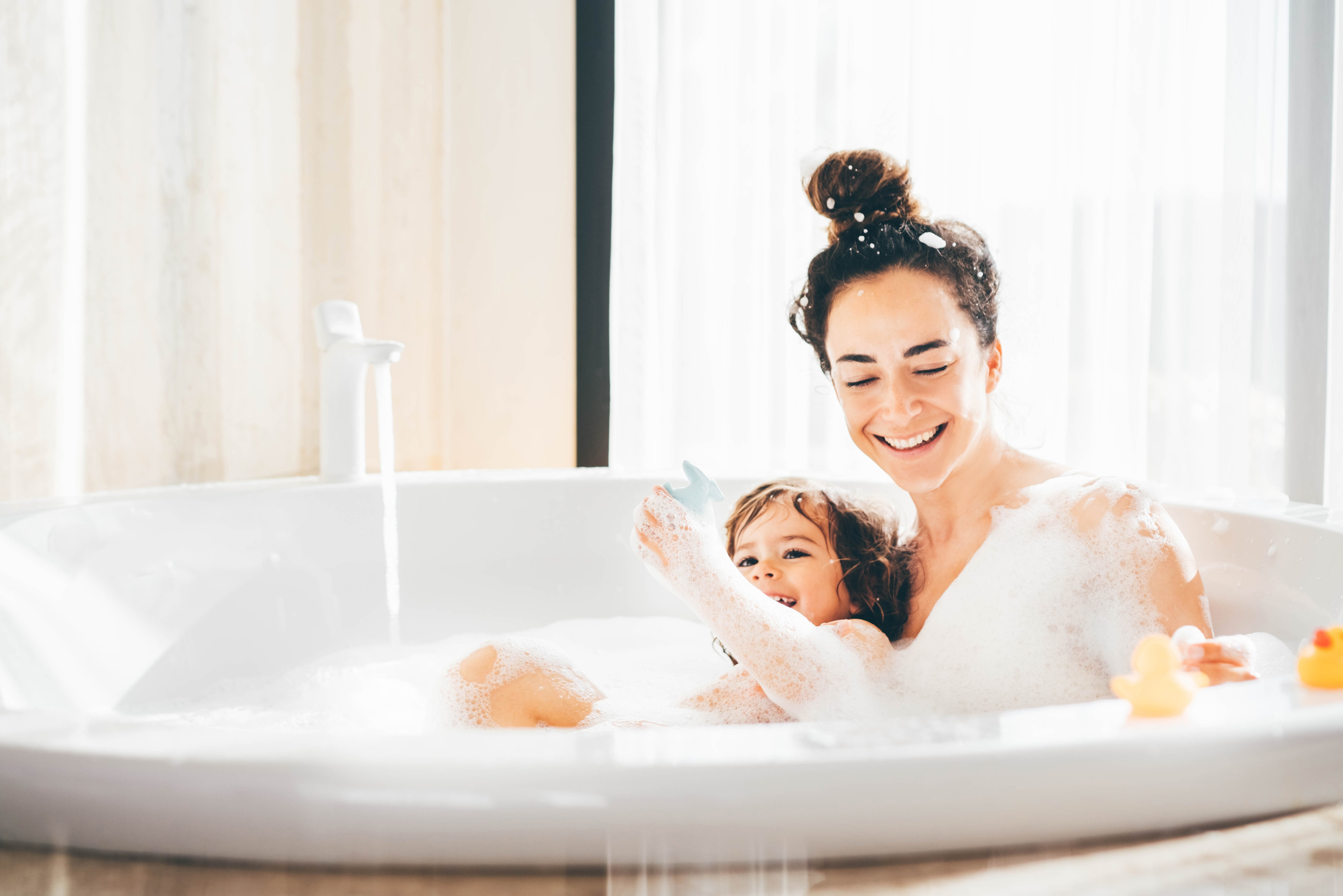 Femme et enfant se relaxant dans un bain moussant avec des canards en caoutchouc sur le bord grâce à l’eau douce de l’adoucisseur d’eau Minimax.