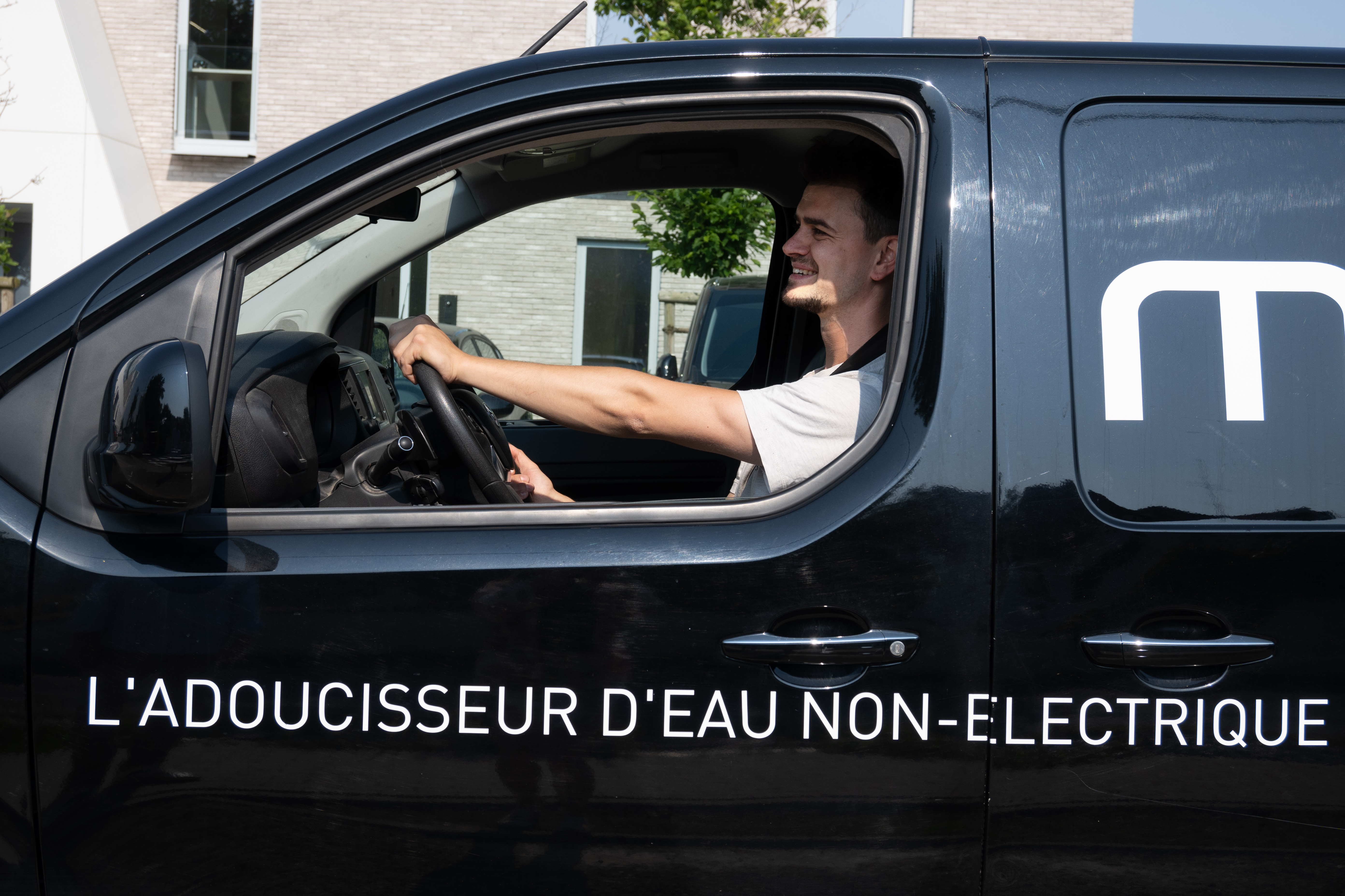 Homme assis en souriant dans une camionnette Minimax avec le texte « L'adoucisseur d'eau non-électrique » sur le côté.