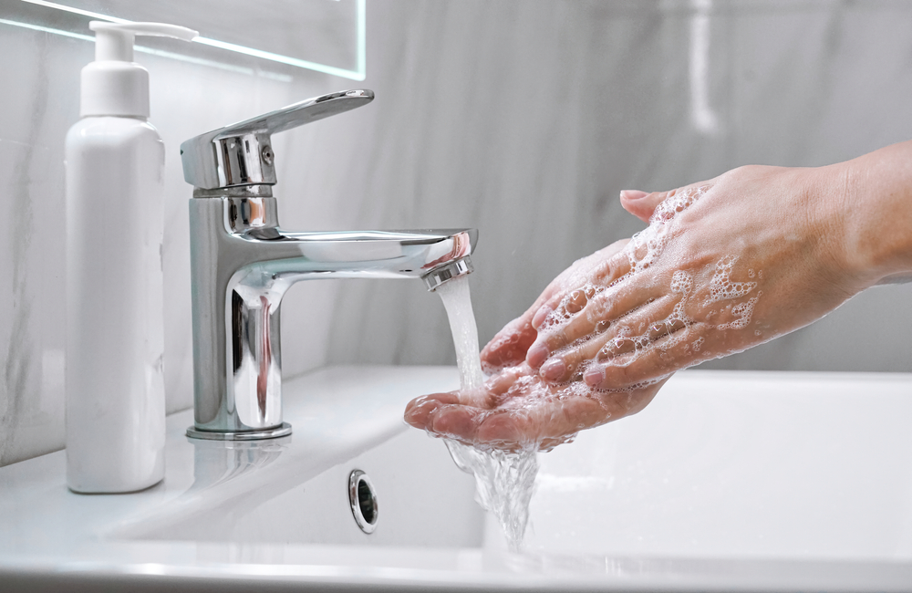 Mains lavées sous un robinet dans une salle de bain avec un distributeur de savon blanc en arrière-plan, avec davantage de mousse et moins de produit grâce à l’adoucisseur d’eau Minimax.