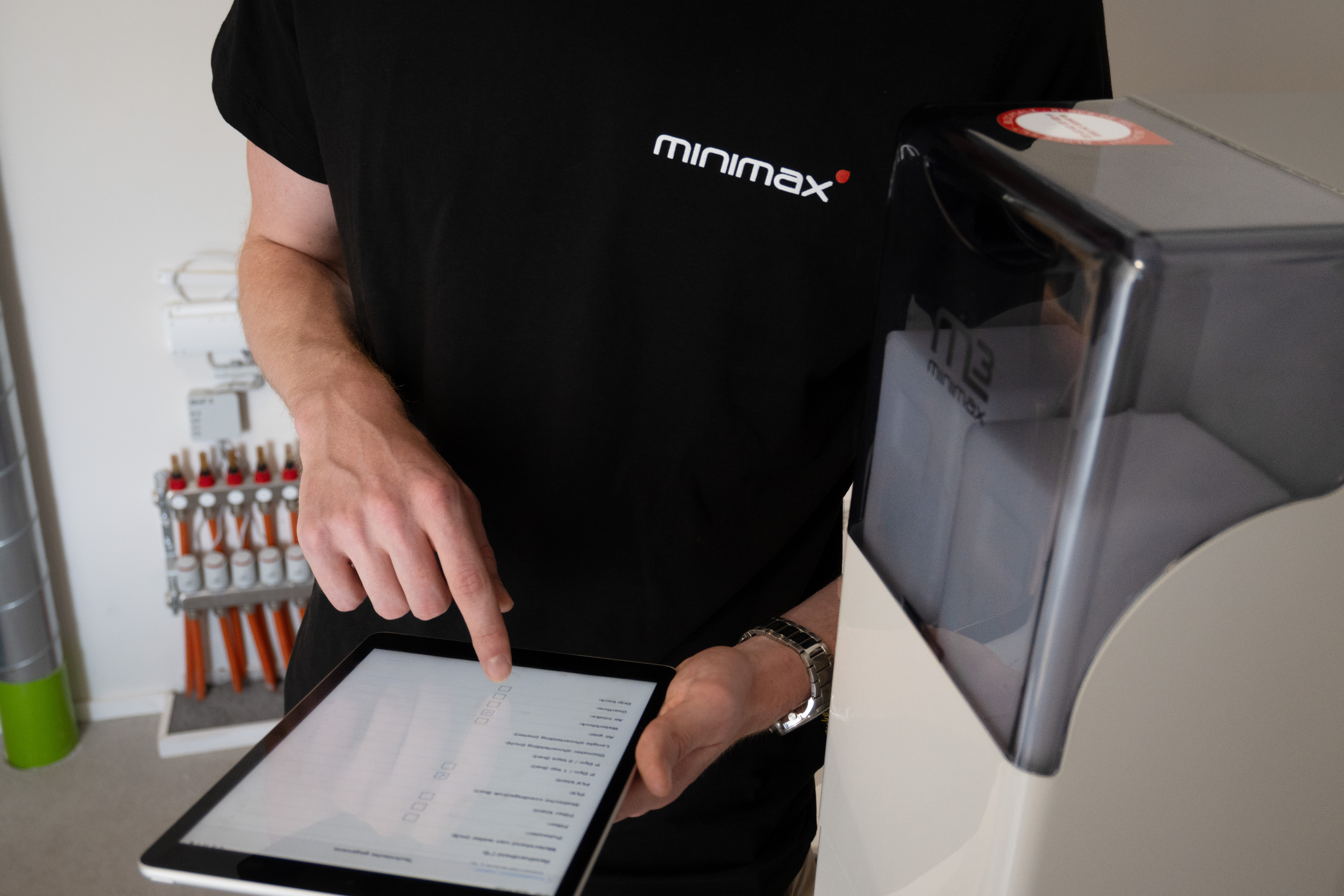 Personne en t-shirt noir utilisant une tablette à côté d’un adoucisseur d’eau Minimax dans un local technique d’une habitation.