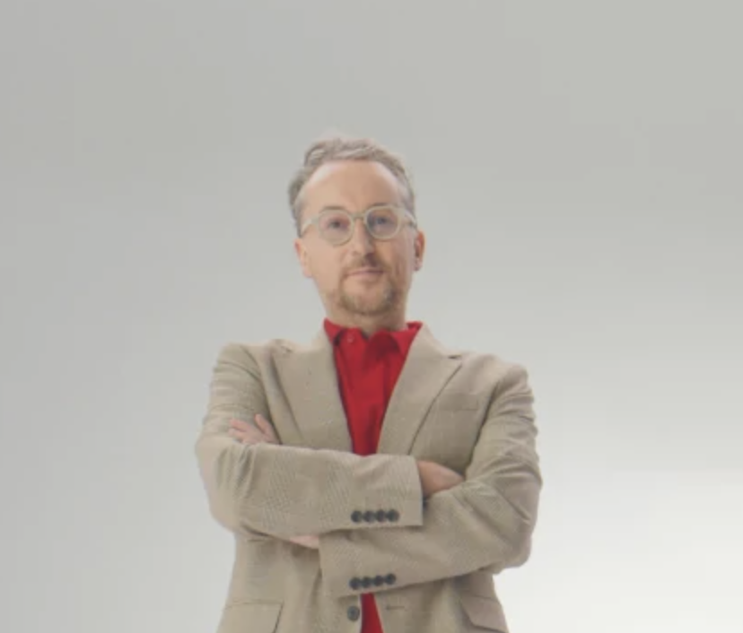 Homme avec lunettes et barbe, portant un blazer beige et une chemise rouge, bras croisés, inventeur de l’adoucisseur d’eau Minimax.