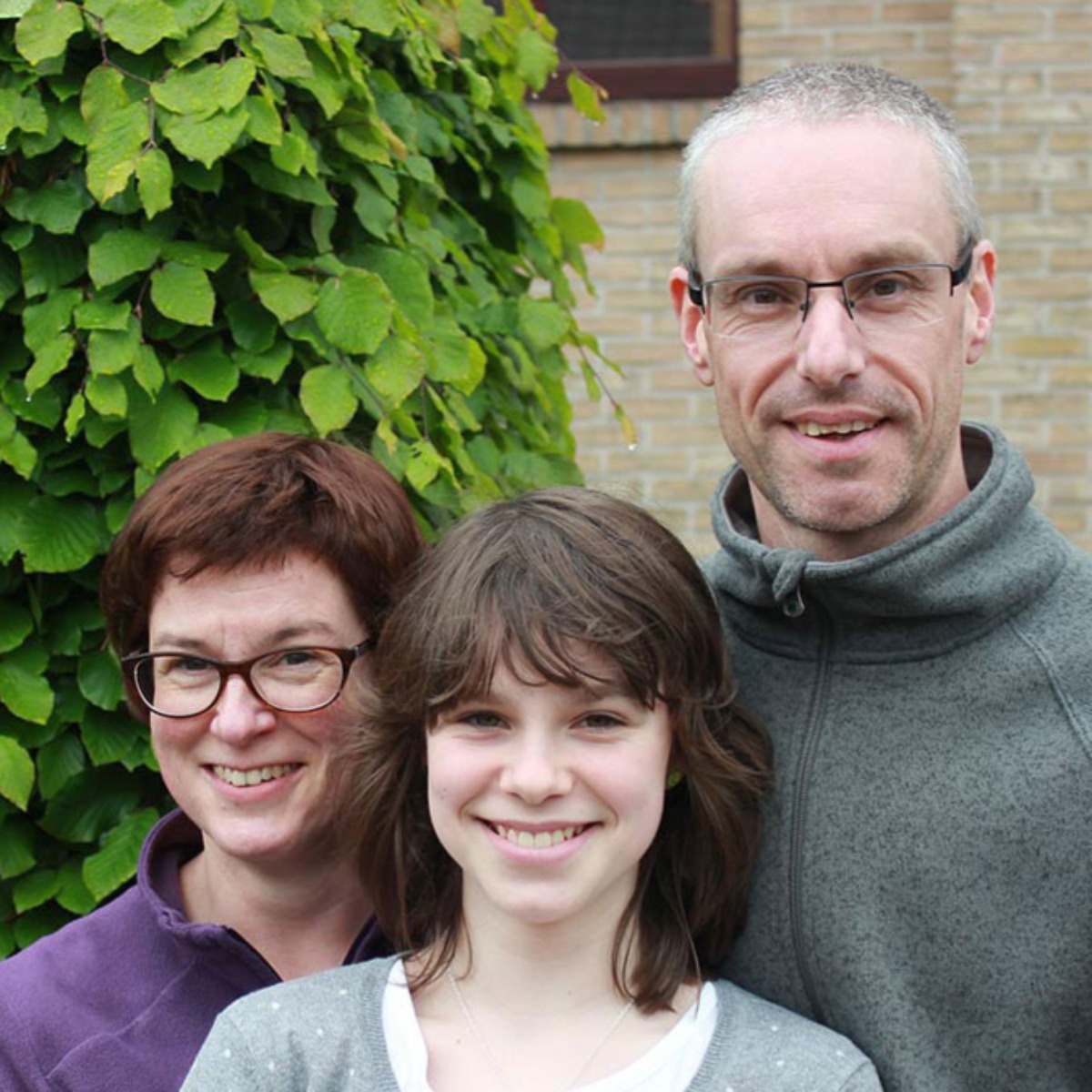 Gezinsfoto met drie personen voor een achtergrond van groene bladeren en een bakstenen muur als tevreden gebruikers van de Minimax waterontharder.